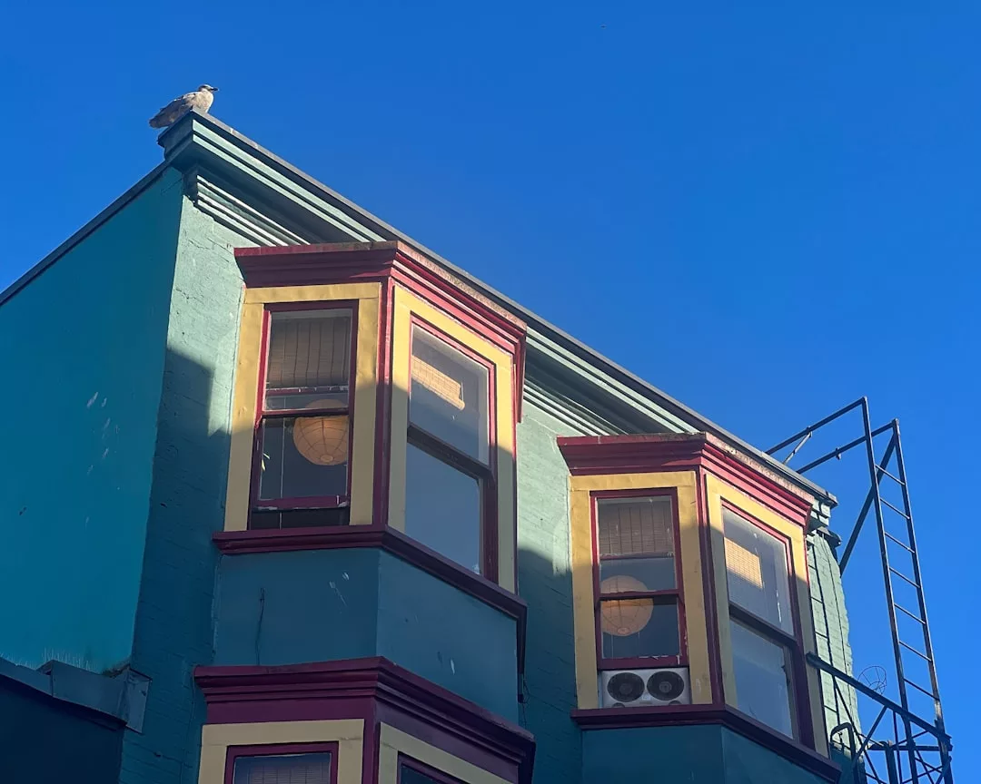 Ein blaugrünes und gelbes Gebäude mit Erkerfenstern und einer Feuertreppe, unter einem klaren blauen Himmel. Ein Vogel sitzt auf dem Dach und unterstreicht die Bedeutung von Immobilienbewertung in einem pulsierenden städtischen Umfeld.