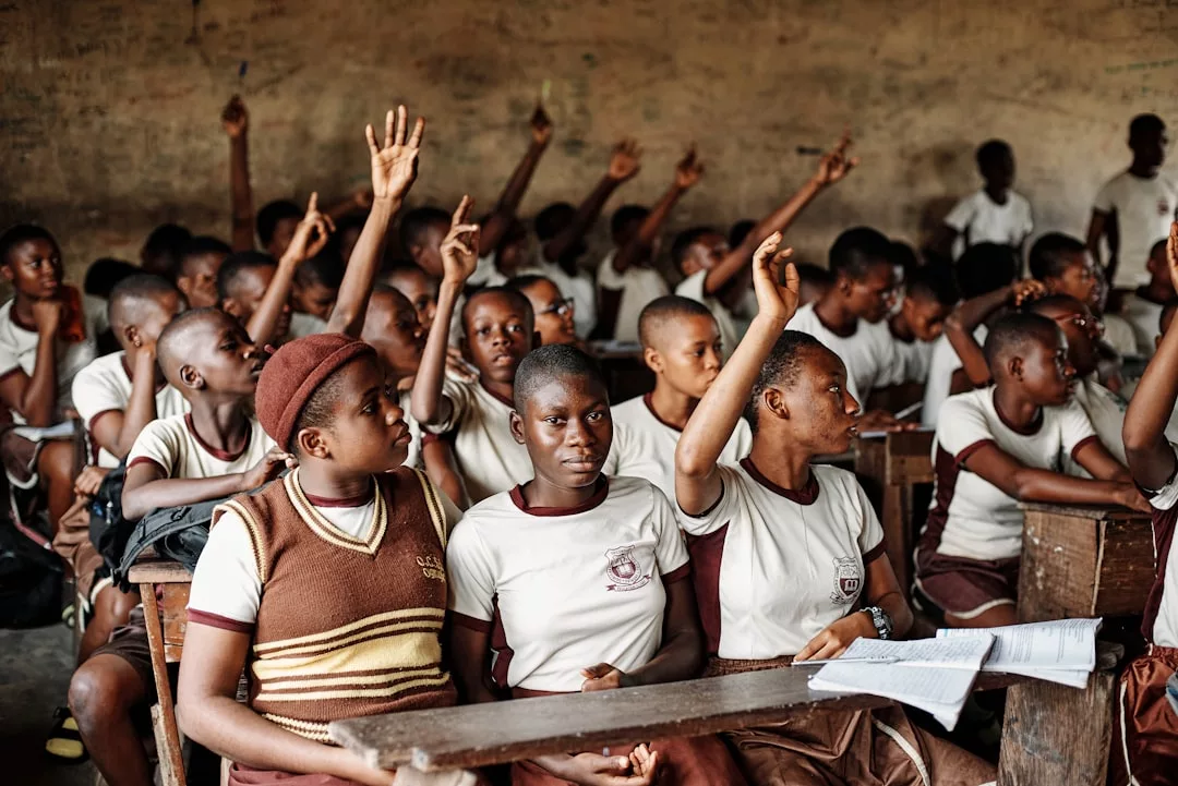 Ein Klassenzimmer mit Kindern in Uniform, von denen einige die Hand heben, um eine Frage zu beantworten, während andere nach vorne oder zur Seite schauen, was ein Gefühl der Gemeinschaft unter den Schülern widerspiegelt.