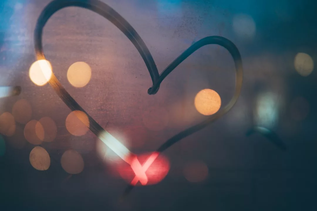 Eine Herzform ist auf ein nebliges Glasfenster mit verschwommenen Lichtern der Stadt im Hintergrund gezeichnet und symbolisiert Liebe und die Wärme zwischenmenschlicher Beziehungen inmitten des städtischen Abends.