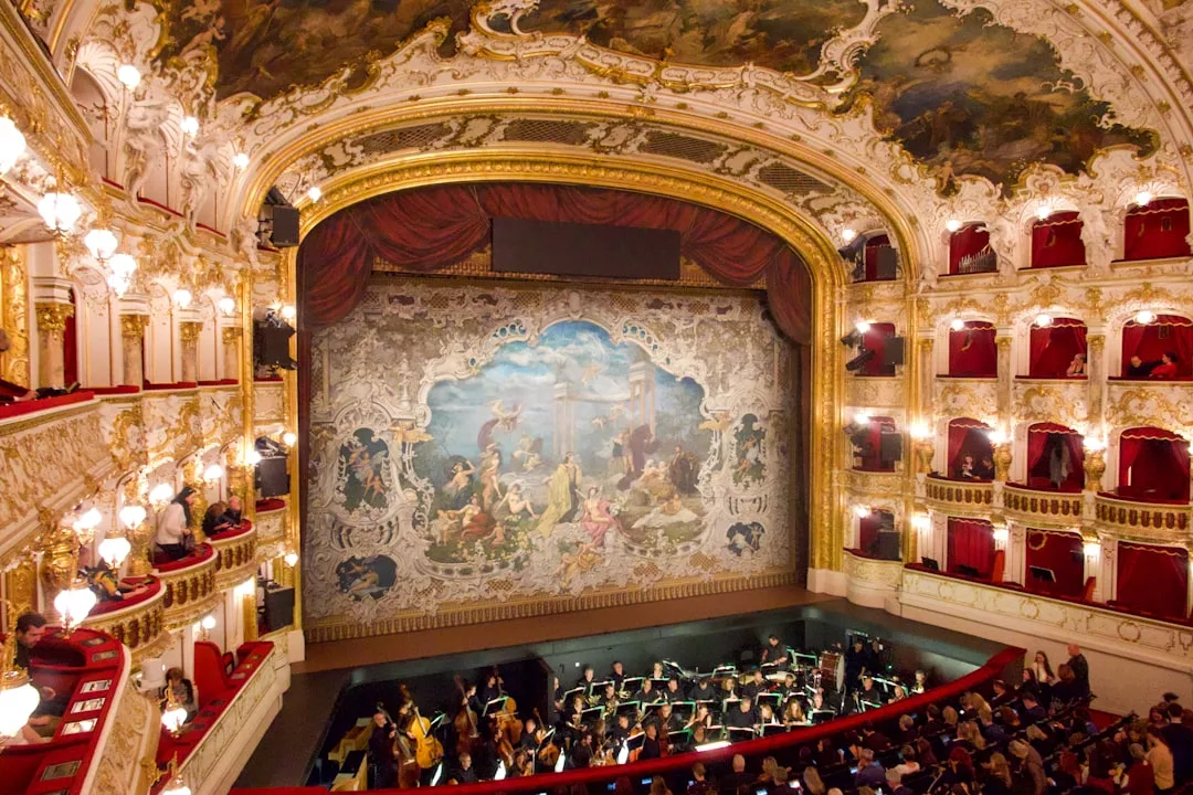 Ausgeschmücktes Theaterinterieur mit Balkonen, einem geschmückten Bühnenvorhang und einem Orchester, das im Orchestergraben spielt - ein Bild für die Bedeutung von Opernaufführungen in der heutigen Gesellschaft.