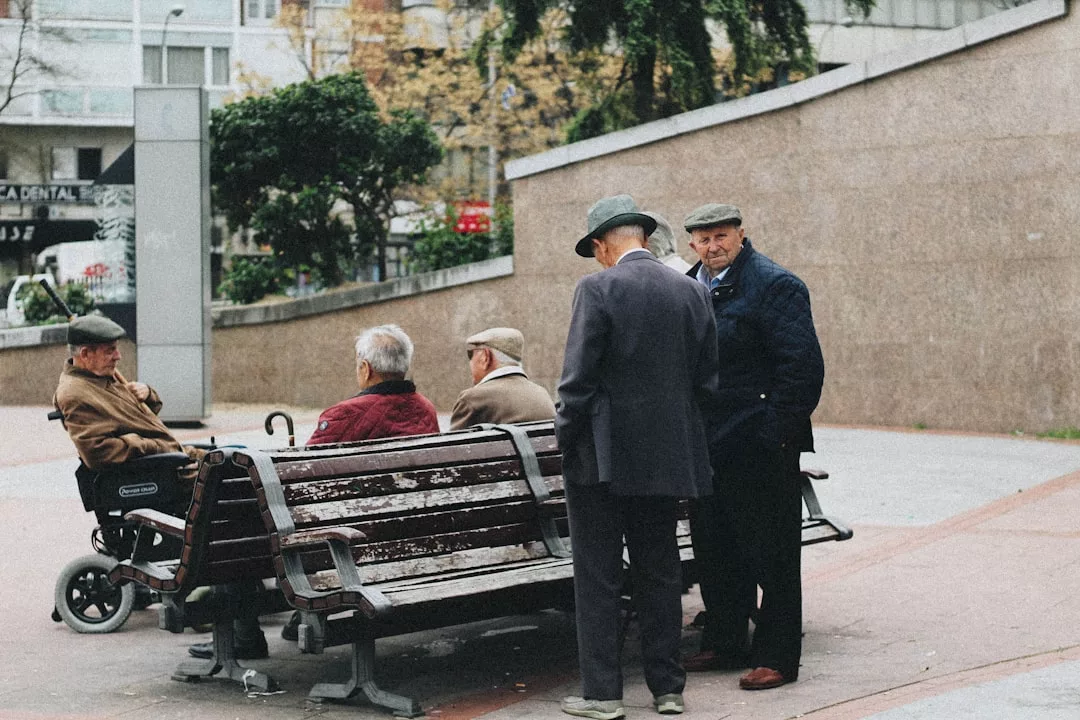 Sechs ältere Männer, teils auf Bänken sitzend, teils stehend, diskutieren in einer städtischen Außenanlage an einer schrägen Betonwand über Themen wie Altersvorsorge.