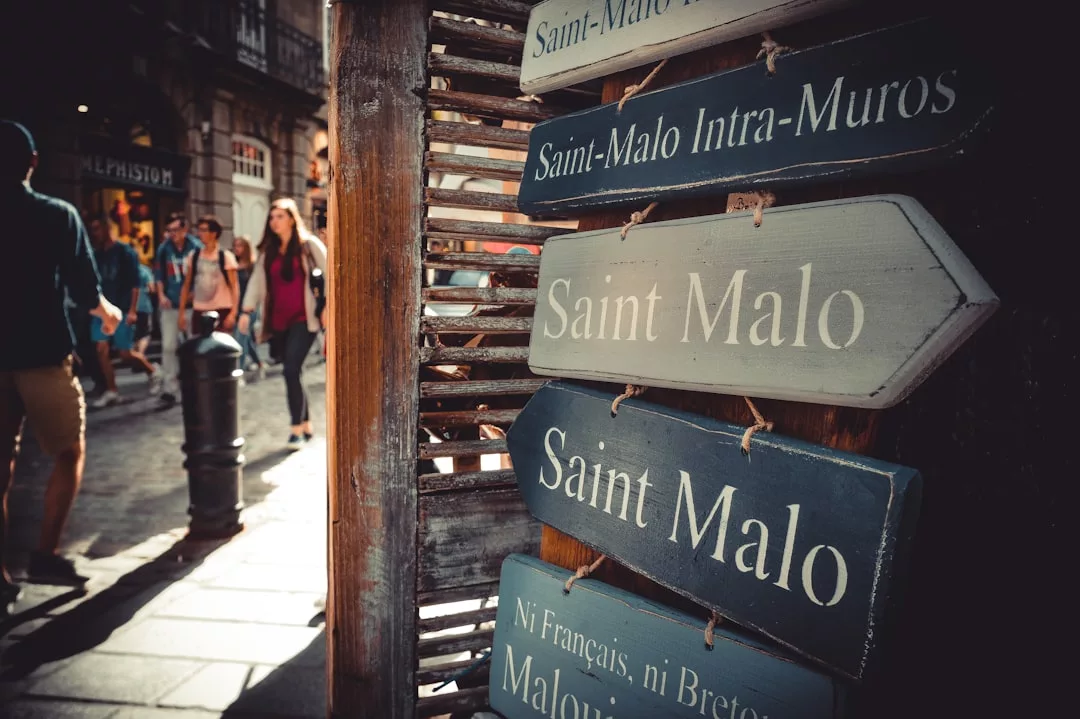 Hölzerne Richtungsschilder weisen an einer belebten Straße mit Spaziergängern im Hintergrund auf "Saint Malo" hin, das in der heutigen Zeit ein Muss ist und oft in Reiseblogs erwähnt wird.