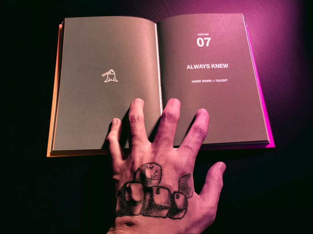 Eine Hand mit einem Würfelmotiv ruht auf einem aufgeschlagenen Buch, das Kapitel 07 mit dem Titel "ALWAYS KNEW: HARD WORK > TALENT" enthält, das die Bedeutung von Selbstliebe und Lebenserfahrungen widerspiegelt.