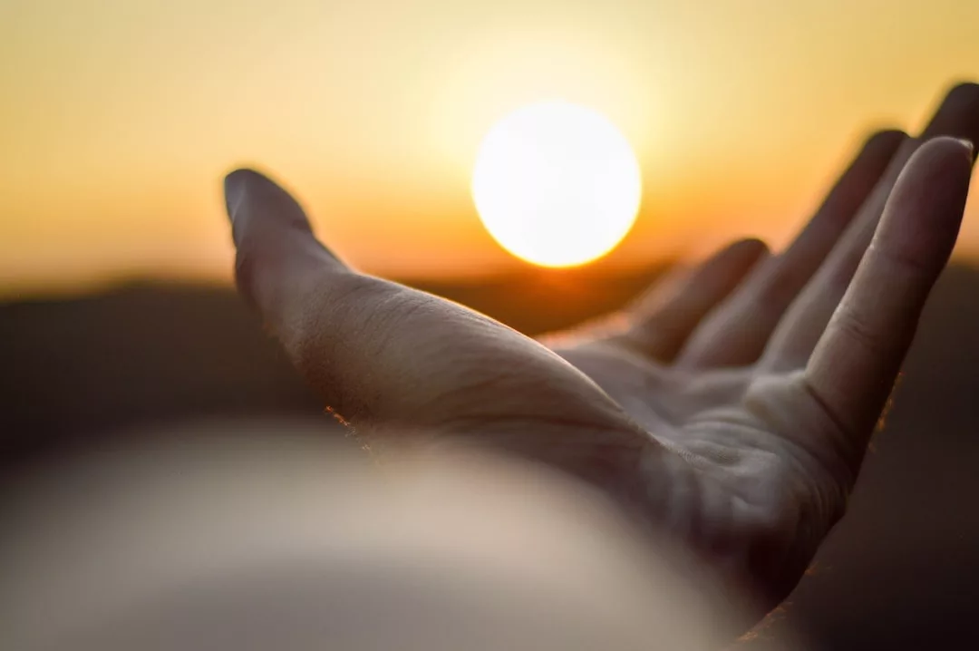 Eine mit der Handfläche nach oben ausgestreckte Hand steht vor einer hellen untergehenden Sonne vor einem verschwommenen Hintergrund und symbolisiert Spiritualität in der modernen Gesellschaft.