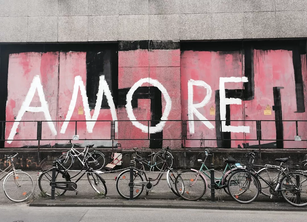 Mehrere Fahrräder stehen auf dem Gehweg vor einer Wand, auf der in großen weißen Buchstaben "AMORE" auf rosa und schwarzem Hintergrund steht. Die Street Art zeigt die Bedeutung von Liebe in der modernen Gesellschaft.