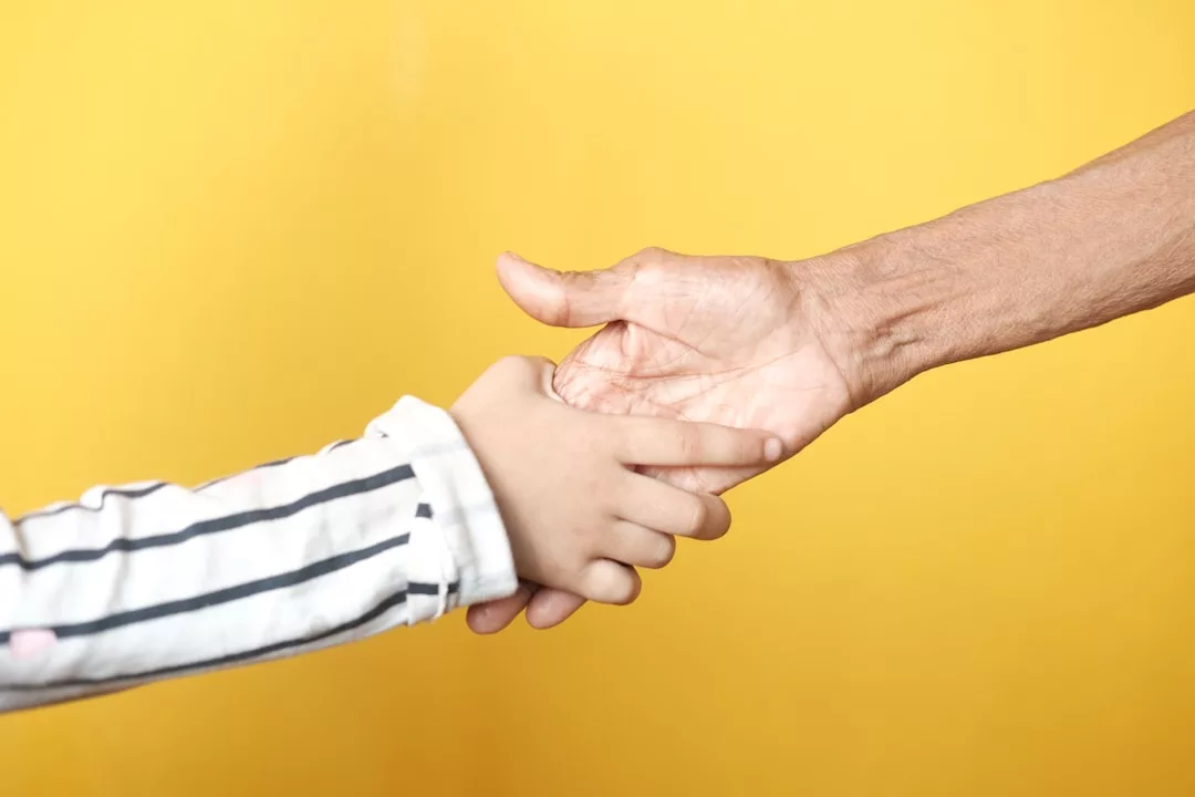 Eine erwachsene Hand, die die Hand eines Kindes hält, vor einem einfarbig gelben Hintergrund, der Vertrauen und die tiefe Bedeutung von unterstützenden Beziehungen symbolisiert.