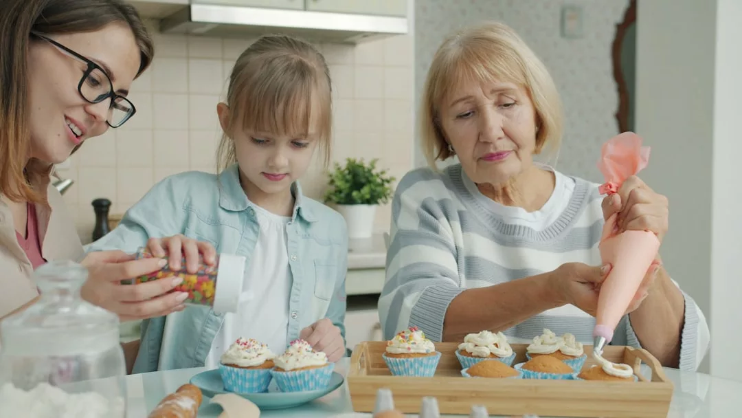Drei Personen verzieren in einer Küche Muffins - eine benutzt einen Spritzbeutel für die Glasur, eine andere streut die Dekoration, und die dritte beobachtet und gibt Tipps für gesunde Ernährung, während sie den kreativen Prozess genießt.