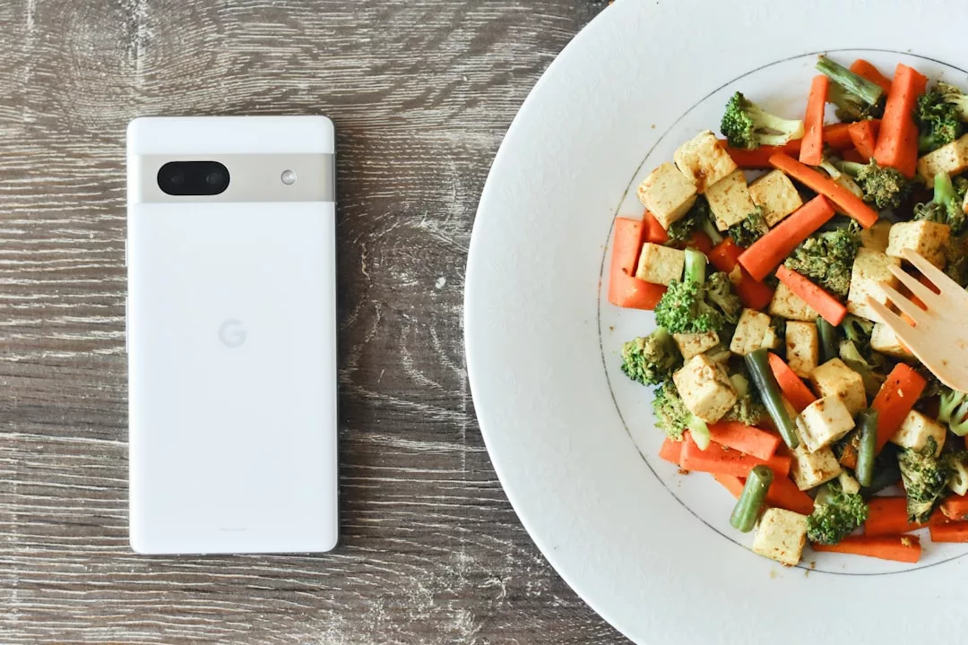 Ein weißes Smartphone liegt auf einer hölzernen Oberfläche neben einem Teller mit Tofu und gemischtem Gemüse mit einer Holzgabel - perfekt, um gesunde Ernährung zu verfolgen oder Ernährungs-Apps zu nutzen.