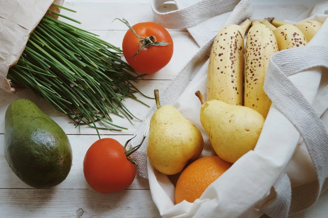 In einer wiederverwendbaren Tasche liegen Bananen, Birnen und eine Orange neben Tomaten, Schnittlauch und einer Avocado auf einer weißen Holzfläche - ideal für Vegetarier, die Ernährungstipps suchen.