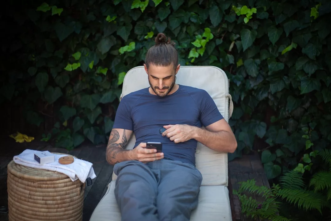 Ein Mann mit Dutt und Tattoo sitzt auf einem Liegestuhl im Freien und surft in Top-Apps oder Fitness-Apps auf seinem Telefon, während im Hintergrund Grünpflanzen wachsen und ein kleiner runder Tisch neben ihm steht.