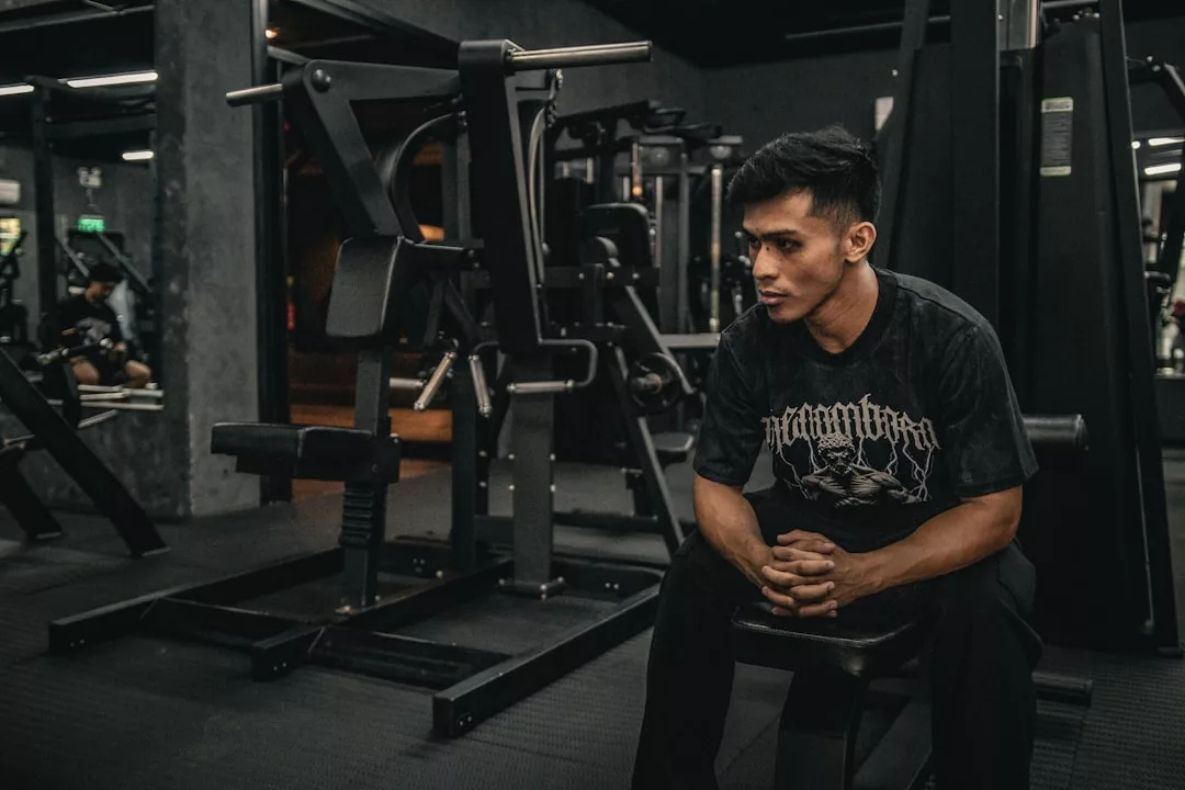 Ein Mann sitzt auf einer Bank in einem schwach beleuchteten Fitnessstudio, faltet seine Hände und blickt nach vorne, umgeben von Kraft- und Trainingsgeräten - vielleicht inspiriert von Fitness-Blogs, ein gesünderes Leben zu führen.