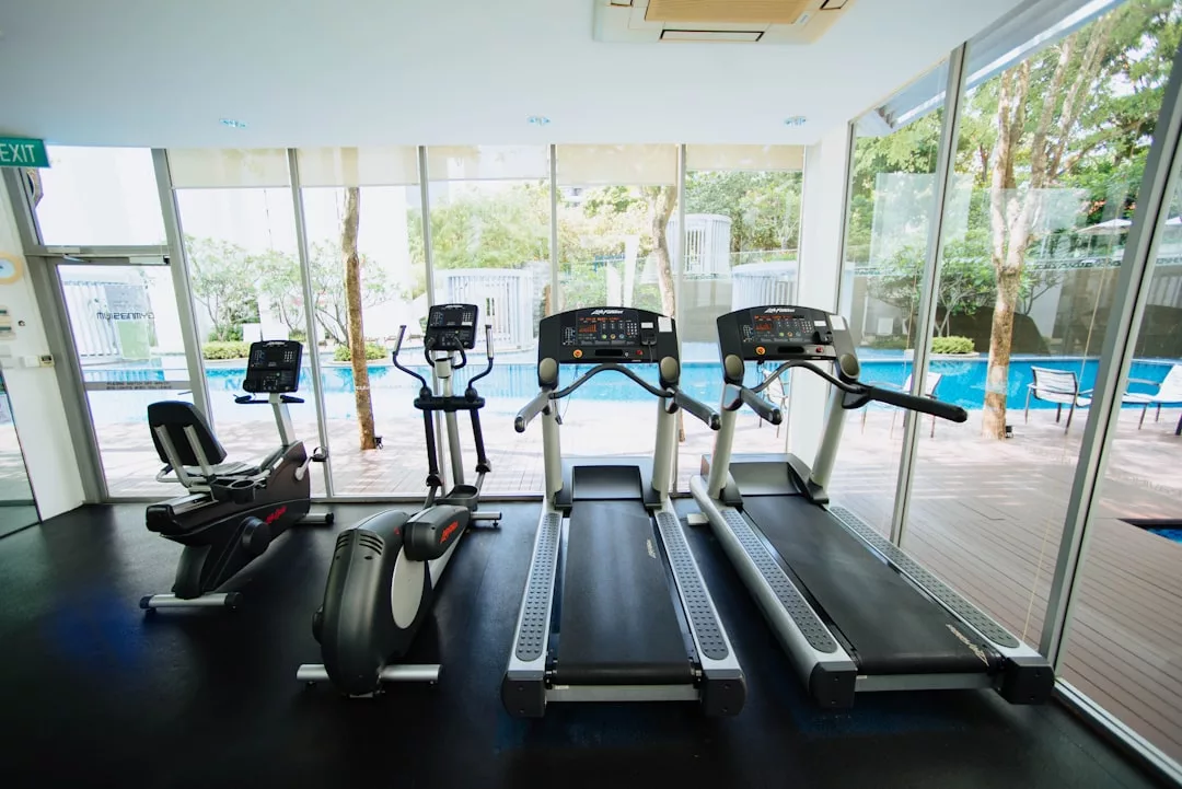 Ein moderner Fitnessraum mit Fitnessgeräten wie zwei Laufbändern, zwei Ellipsentrainern und einem Heimtrainer, perfekt für effektives Training, während man durch große Fenster den Blick auf den Pool und den Außenbereich genießen kann.