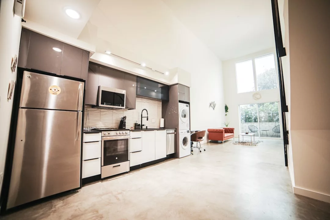 Moderne offene Küche mit hochwertigen Haushaltsgeräten, Waschmaschine und Trockner, sowie ein Wohnbereich mit rotem Sofa und großen Fenstern im Hintergrund für effizientes Wohnen.