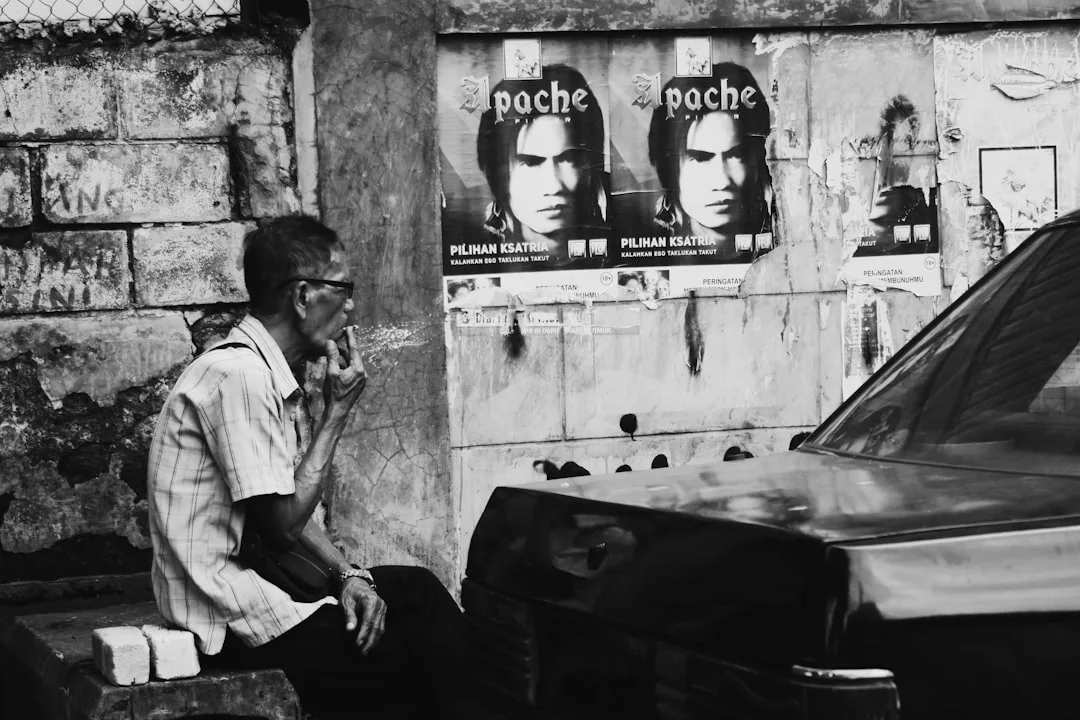 Ein älterer Mann sitzt rauchend auf einer Bank neben einer mit Plakaten bedeckten Wand, während ein geparktes Auto und das Echo des Jazz auf die musikalische Geschichte und Entwicklung dieses pulsierenden städtischen Umfelds hinweisen.