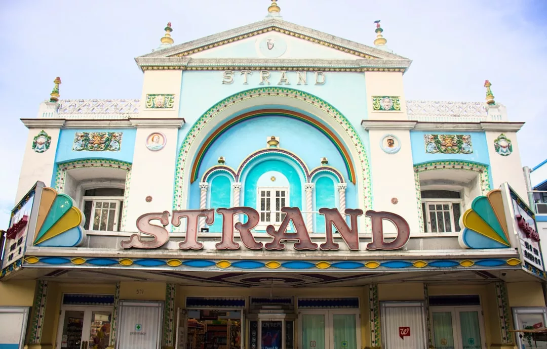 Art-Deco-Theaterfassade mit pastellblauen und weißen Farben, dekorativen Details, die antike Einflüsse widerspiegeln, und großen Buchstaben über dem Eingang, die "STRAND" bedeuten.
