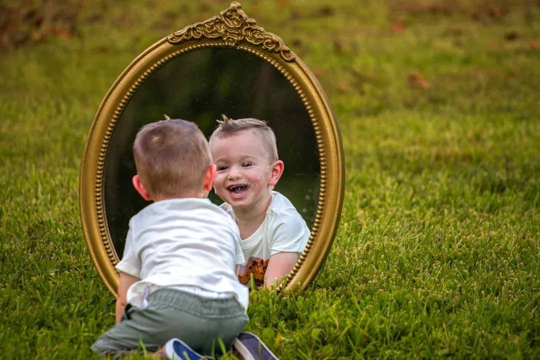Ein kleines Kind inmitten von Babys Entwicklung kniet im Gras vor einem großen, verzierten Spiegel und lächelt sein Spiegelbild an.