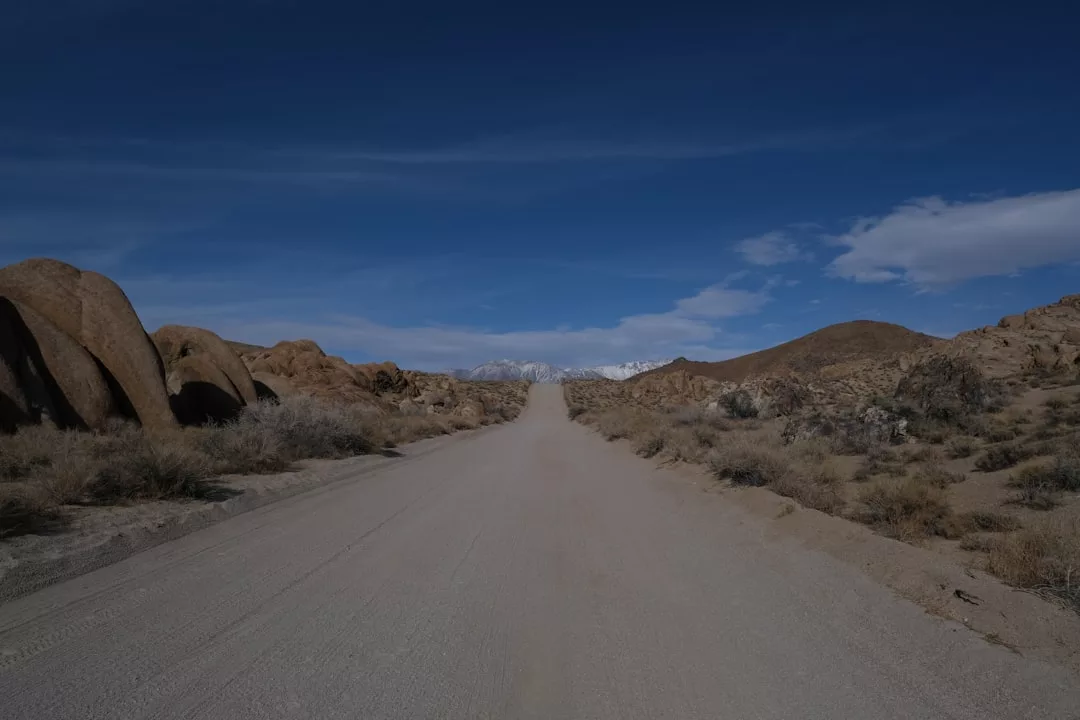 Eine gerade Schotterstraße führt durch eine trockene, felsige Wüstenlandschaft unter einem klaren, blauen Himmel mit schneebedeckten Bergen in der Ferne - perfekt für Roadtrips auf vier Rädern und alle, die die Welt entdecken möchten.