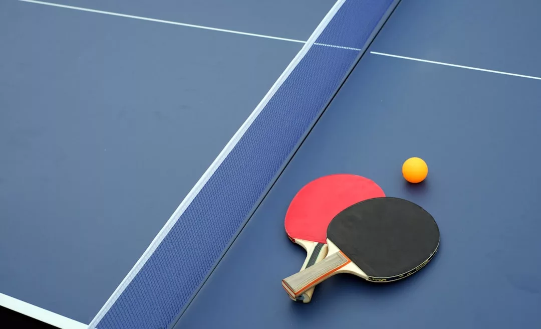 Zwei Tischtennisschläger und ein orangefarbener Ball liegen auf einem blauen Tisch in der Nähe des weißen Netzes, bereit für ein lustiges Sportspiel, das für alle Altersgruppen geeignet ist.