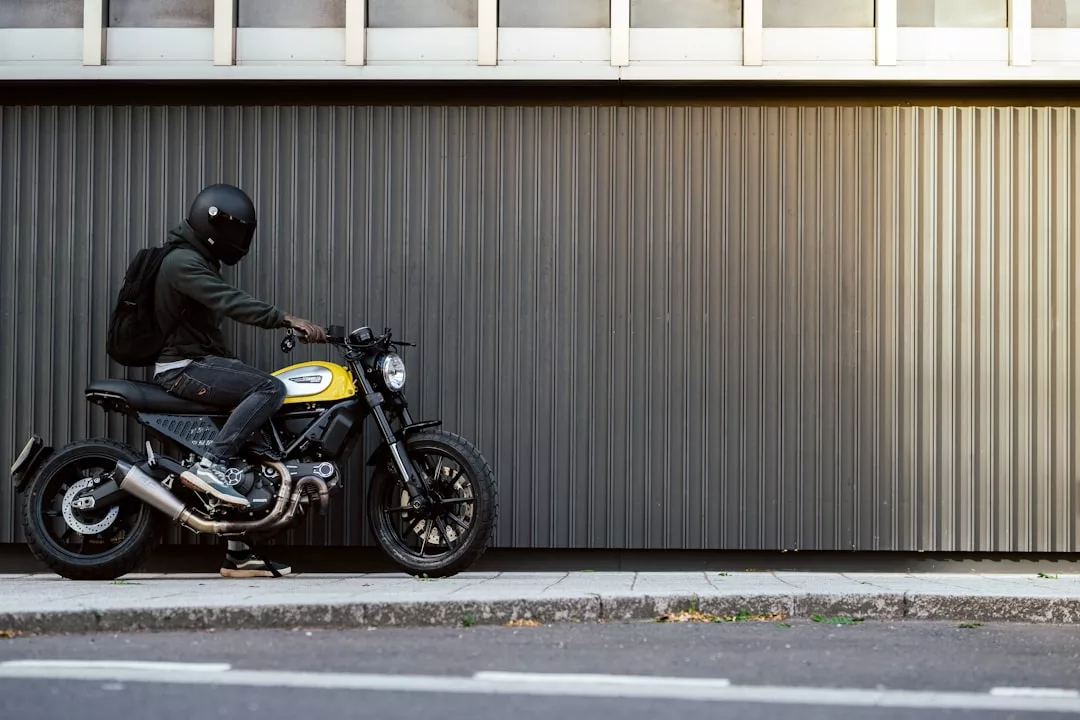 Eine Person mit Helm und Rucksack sitzt auf einem gelb-schwarzen Motorrad, das auf dem Gehweg vor einer großen grauen Metalltür geparkt ist und die Freude am Motorradfahren und zeitlosen Genuss verkörpert.