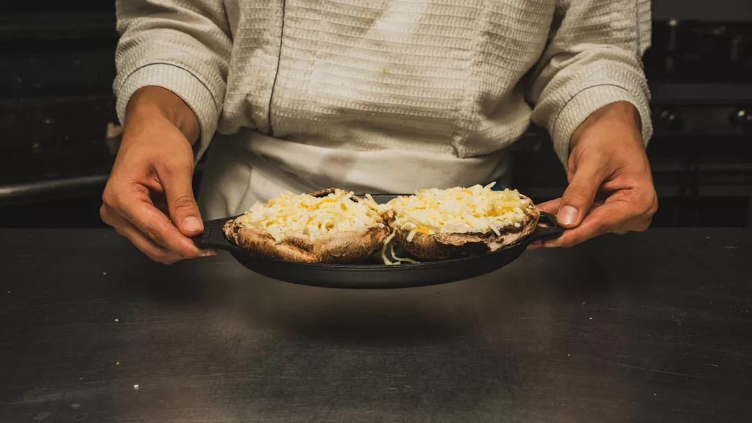 Eine Person im Kochmantel hält eine Pfanne mit zwei großen Pilzen, die mit geriebenem Käse überbacken sind und die Grundlagen der italienischen Küche darstellen.