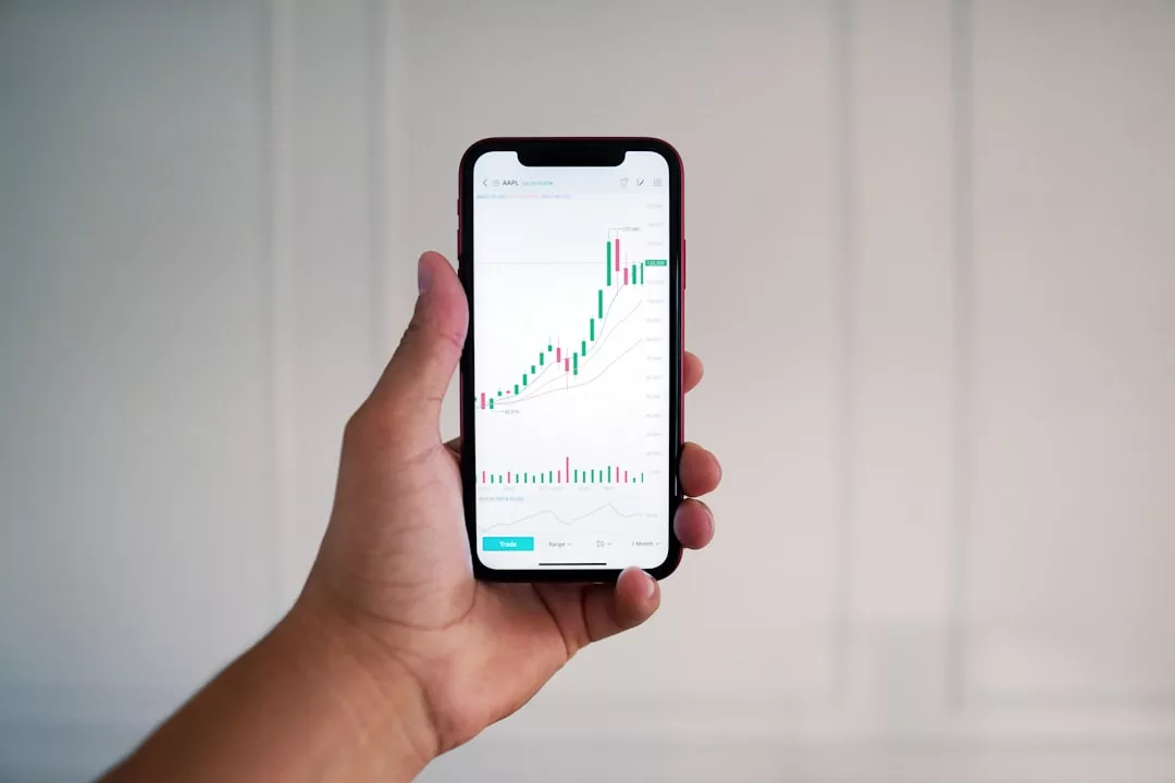 Eine Hand, die ein Smartphone hält, zeigt ein Finanzdiagramm mit steigenden grünen und roten Kerzenständern und Volumenbalken, das sich perfekt zur Veranschaulichung der Grundlagen des Investierens oder als Teil eines Investieren-Leitfadens eignet.