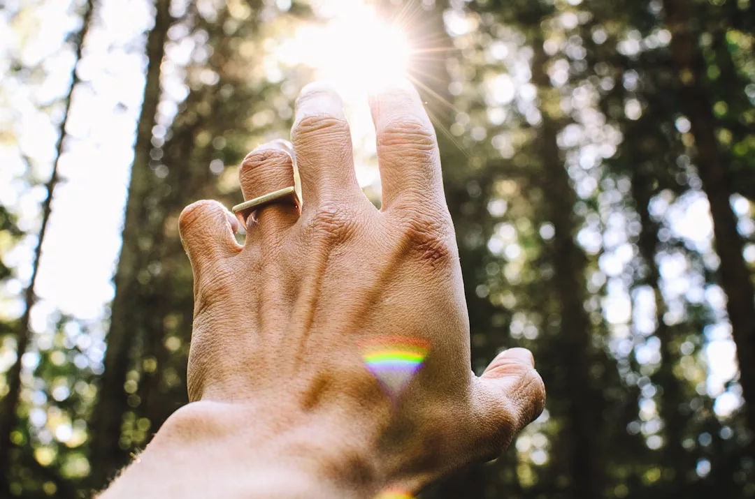 Eine Hand mit einem Ring streckt sich in einem Wald dem Sonnenlicht entgegen, wobei Sonnenstrahlen und ein kleiner Regenbogen zu sehen sind, der an die heilende Kraft der Natur und die sanfte Berührung der Reiki-Heilung erinnert.