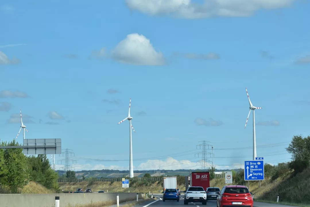Autos fahren auf einer Autobahn mit Windturbinen, die für Windenergie stehen, Stromleitungen und einem Straßenschild im Hintergrund unter einem blauen Himmel mit vereinzelten Wolken.