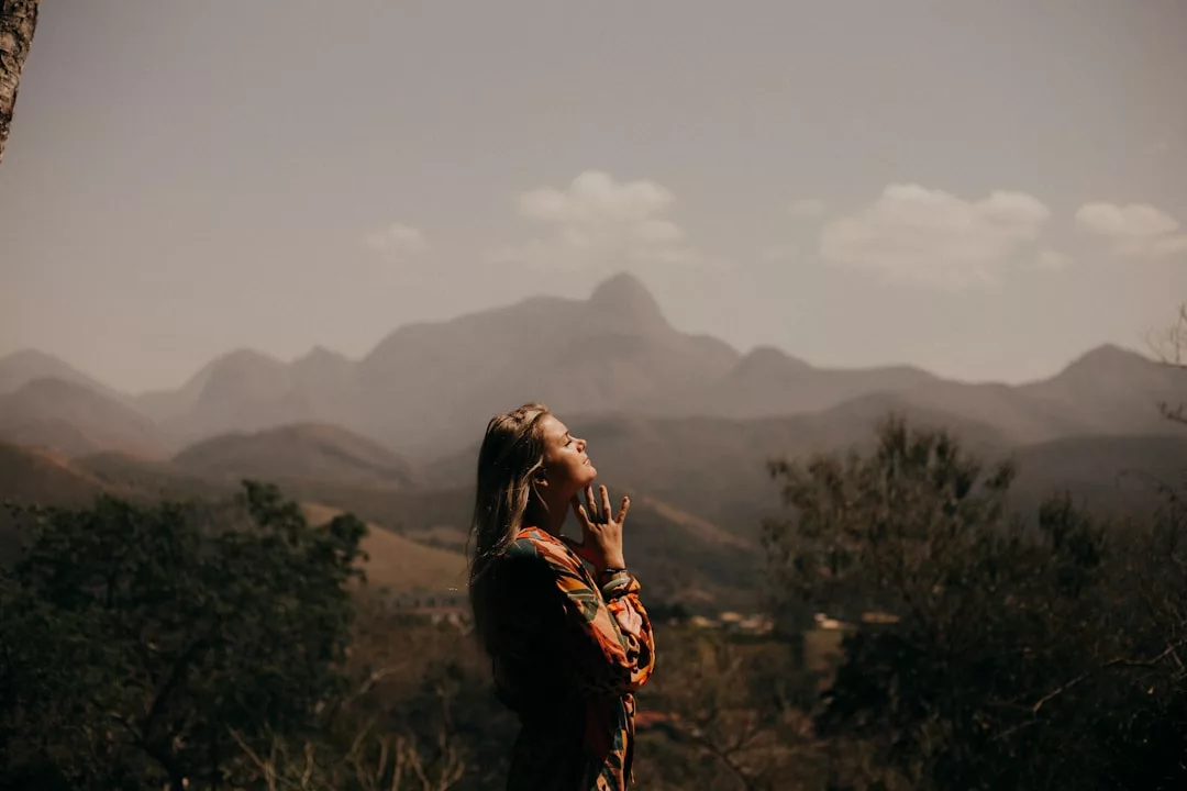 Eine Frau steht mit geschlossenen Augen und gefalteten Händen im Freien, umarmt das Sonnenlicht und die Affirmationen für positive Gedanken, mit Bergen und Bäumen im Hintergrund.