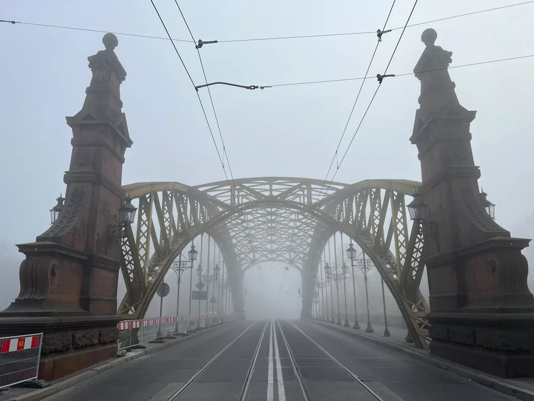 Ein nebliger Blick auf eine große, verzierte Metallbrücke mit steinernen Pfeilern, Straßenbahnschienen und Oberleitungen, mit eingeschränkter Sicht aufgrund des Nebels - das fängt die Mystik ein, die man oft in inspirierenden Reisevideos findet.