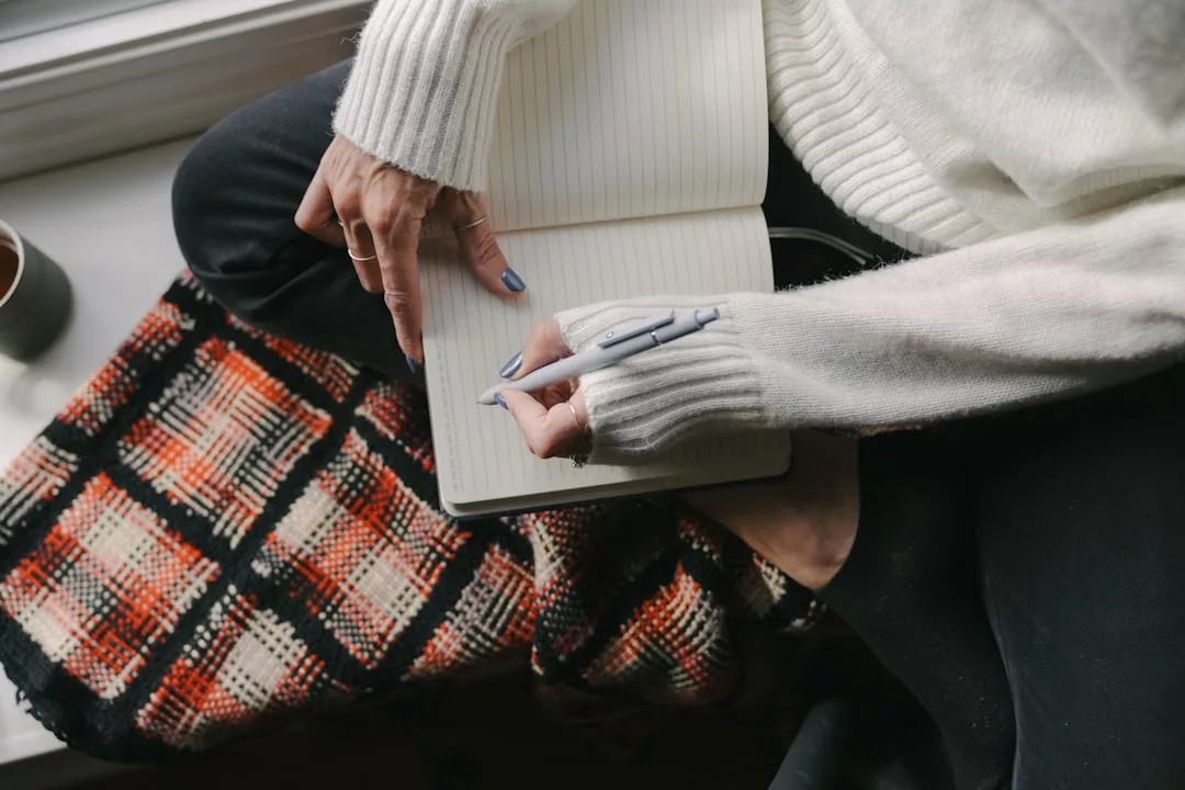 Eine Person in einem weißen Pullover sitzt im Schneidersitz auf einer karierten Decke, hält einen Stift in der Hand und schreibt in ein offenes, leeres Notizbuch - vielleicht beginnt sie ihren persönlichen Leitfaden zur Selbstheilung.