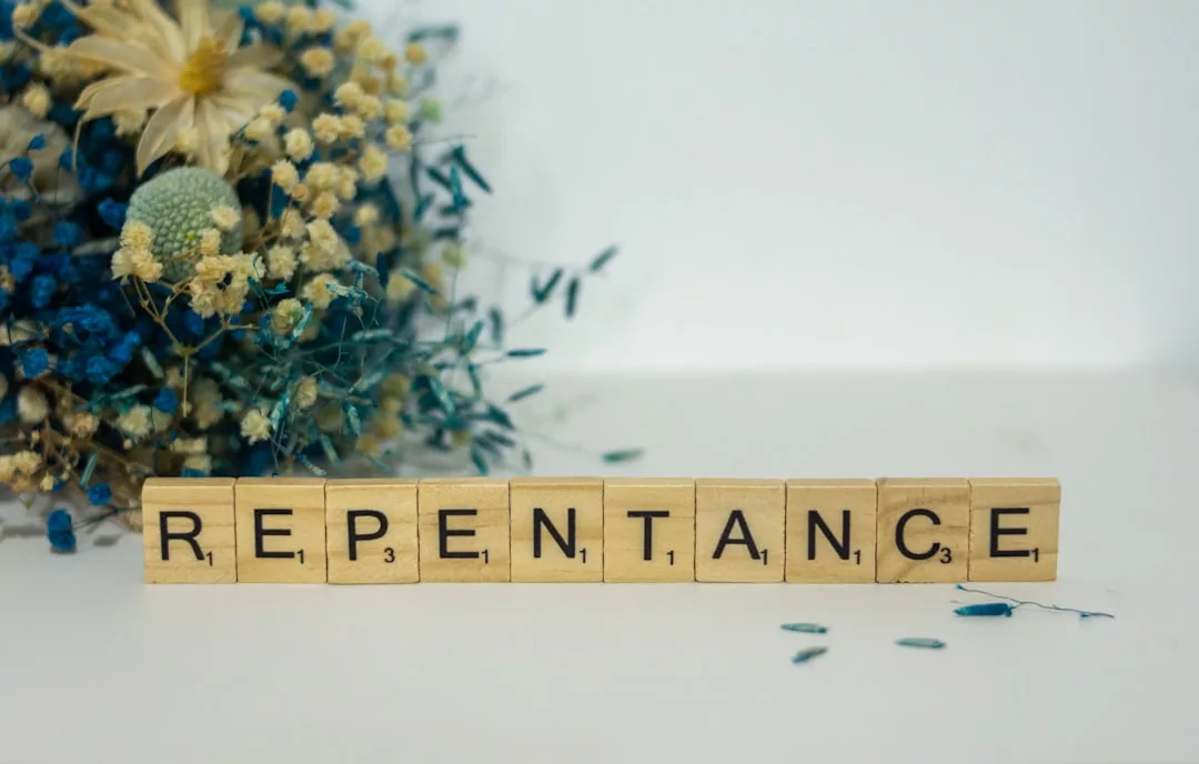 Die hölzernen Scrabble-Kacheln buchstabieren "REPENTANCE" auf einer weißen Fläche, mit getrockneten blauen und gelben Blumen auf der linken Seite, die an die Kraft der Vergebung und die sanfte Schönheit der Vergebung erinnern.