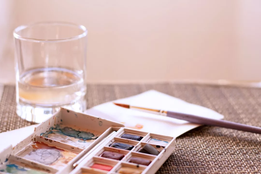 Ein Glas Wasser, ein Aquarellmal-Set, ein Pinsel und ein Blatt Papier werden auf eine gewebte Fläche gestellt - perfekt für erste Tipps und Techniken der Aquarellmalerei.