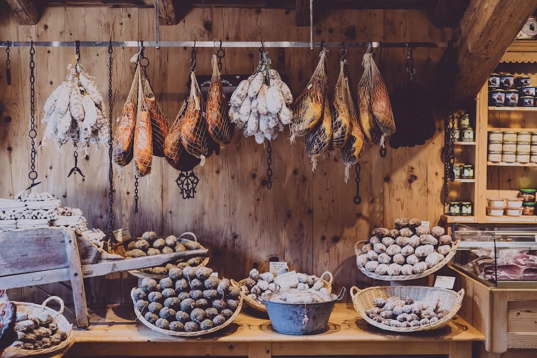 Verschiedene Wurstsorten hängen über Körben mit verschiedenen Wurstsorten und Salami, die traditionelle Zubereitungen in einer charmanten Holztheke in einem rustikalen Laden präsentieren.