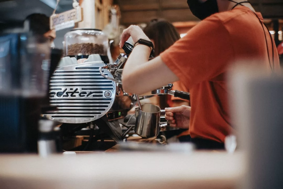 Der Barista im orangefarbenen Hemd demonstriert die Kaffeezubereitung und gibt Kaffee-Tipps, während er ein Kaffeegetränk mit einer professionellen Espressomaschine in einer Caféumgebung zubereitet.