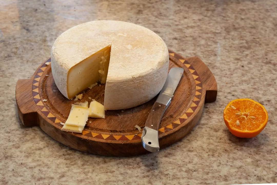 Ein runder Laib Käse mit einem ausgeschnittenen Keil liegt auf einem Holzbrett mit einem Messer, daneben liegen Käsescheiben und eine halbe Orange, die die Kunst der Käseherstellung zeigen.