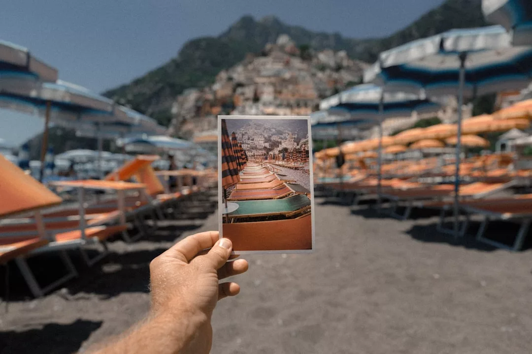 Eine Hand hält ein Foto der gleichen Strandszene, die gerade betrachtet wird und Reihen von orangefarbenen Liegestühlen und Sonnenschirmen mit einer Stadt am Hang im Hintergrund zeigt - perfekt für Reisefotografie und atemberaubende Bilder.