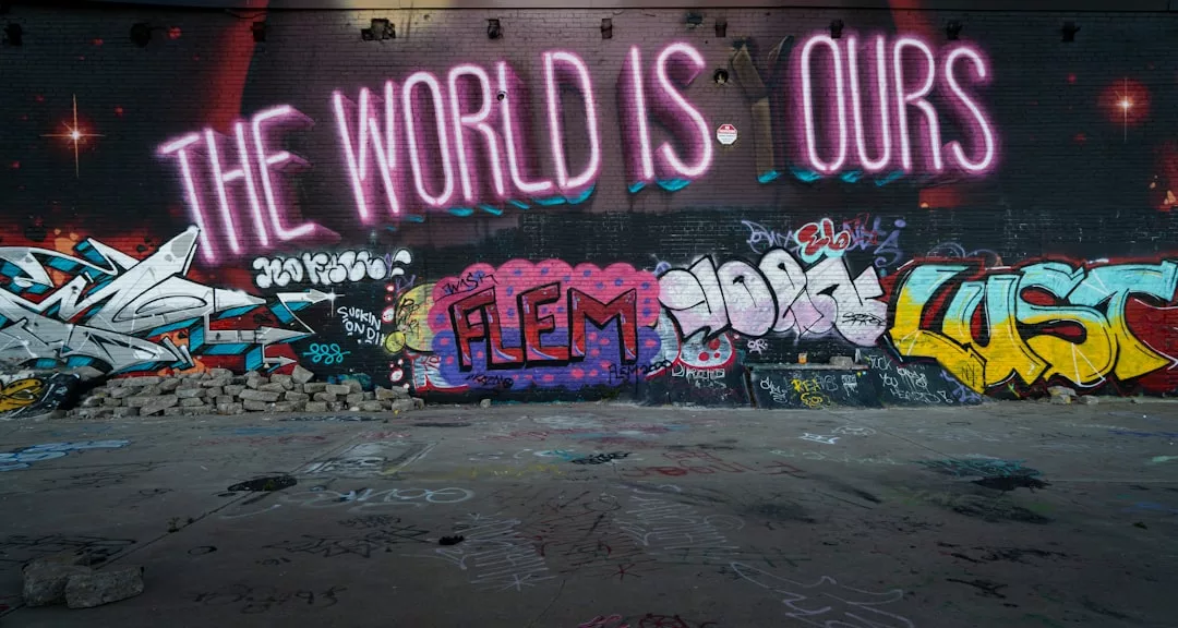 An einer mit Graffiti bedeckten Wand ist ein großer Neon-Schriftzug mit der Aufschrift "THE WORLD IS OURS" über verschiedenen bunten Anhängern und lebendiger Street Art zu sehen, die die urbane Kunst feiert.