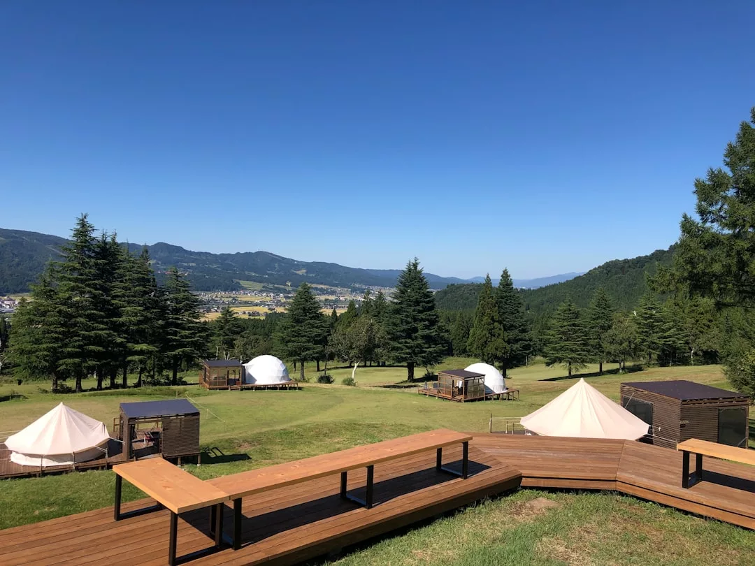 Eine grasbewachsene Wiese mit Holzbänken, Zelten, kleinen Holzhütten und Bäumen bietet einen Hauch von Glamping, eingebettet in die Hügel unter einem klaren blauen Himmel - eine perfekte Mischung aus Natur und Komfort.