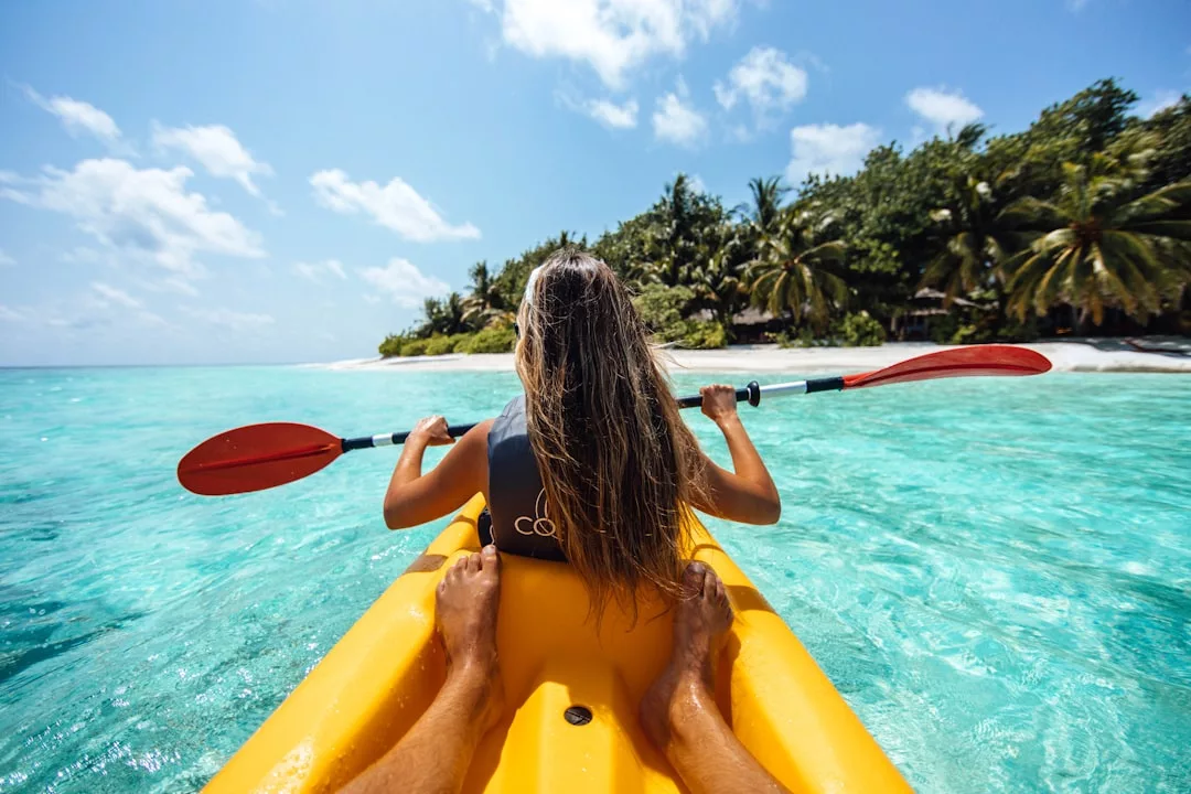 Eine Person mit langen Haaren paddelt in einem gelben Kajak auf klarem türkisfarbenem Wasser in Richtung eines tropischen Strandes mit Palmen unter blauem Himmel und stellt Kajakfahren Techniken vor.