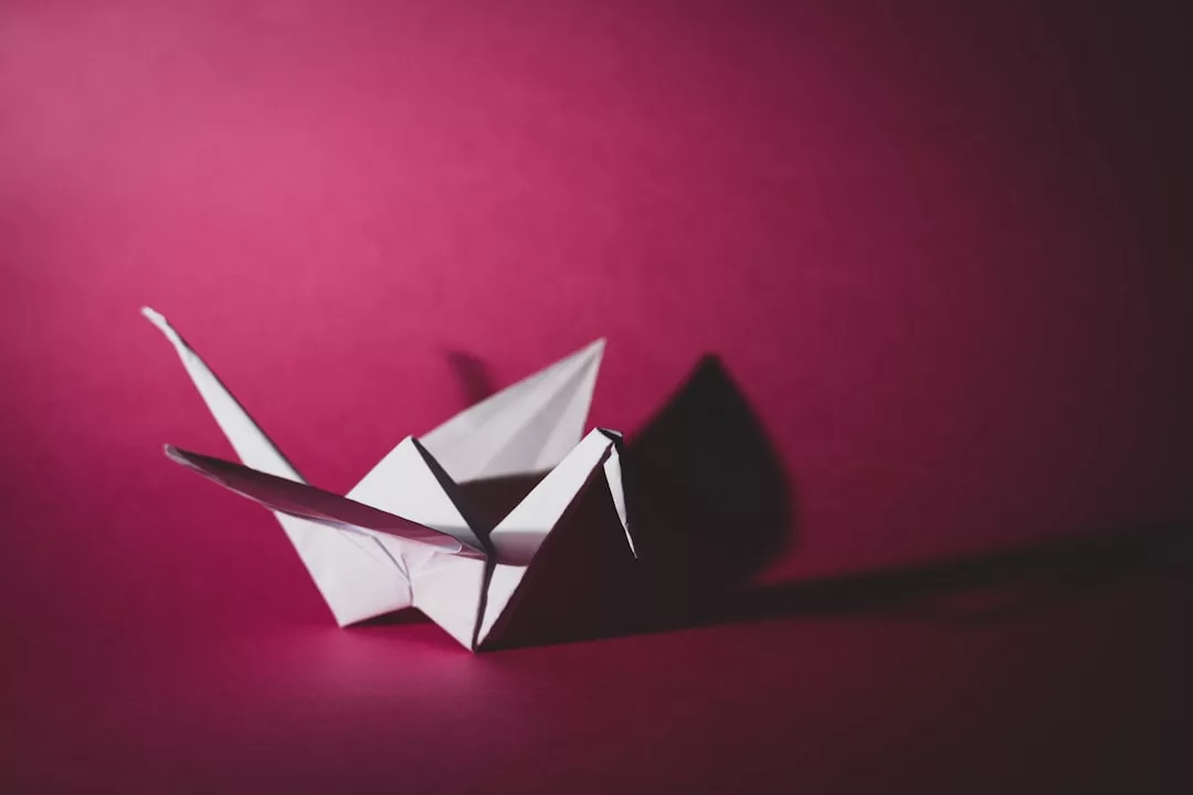 Ein weißer Origamipapierkranich sitzt auf einer Fläche vor einem einfarbigen magentafarbenen Hintergrund und zelebriert die Tradition dieser zarten Kunst, wobei die Schatten nach rechts fallen.
