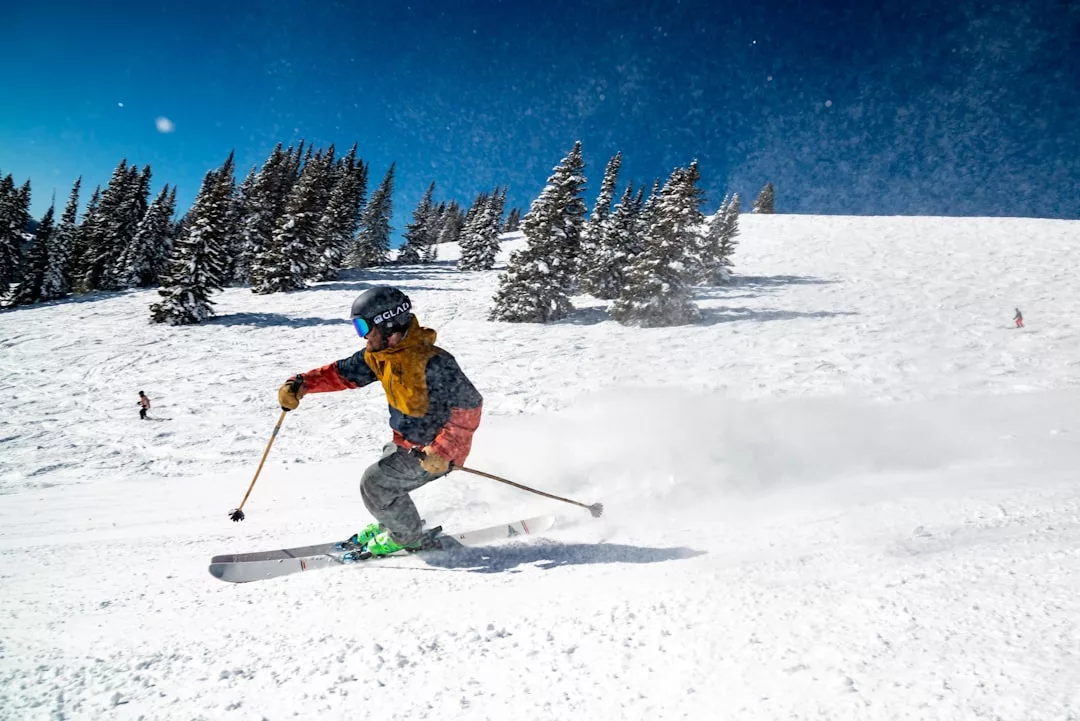 Ein Skifahrer, der einen Helm und eine Skibrille trägt, fährt auf einer verschneiten Piste mit Kiefern und klarem blauen Himmel im Hintergrund bergab. Er demonstriert Skifahren Techniken und genießt Tipps für eine bessere Leistung.