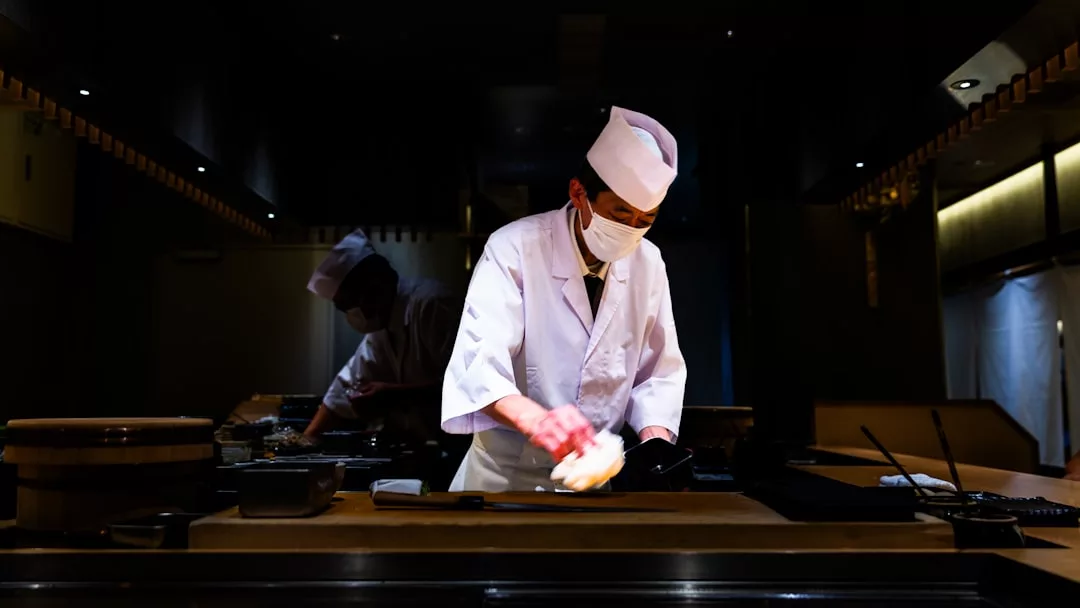 Ein Koch in weißer Uniform und mit Gesichtsmaske bereitet Sushi mit Präzision an einem Tresen in einer schwach beleuchteten Restaurantküche zu. Ein anderer Koch arbeitet im Hintergrund und hält die Tradition aufrecht.