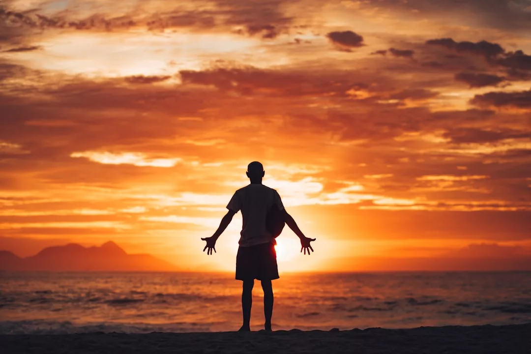 Eine Person steht bei Sonnenuntergang mit ausgestreckten Armen am Strand, blickt auf den Ozean und einen dramatischen orangefarbenen Himmel, strahlt Dankbarkeit aus und umarmt das Glück in diesem ruhigen Moment.