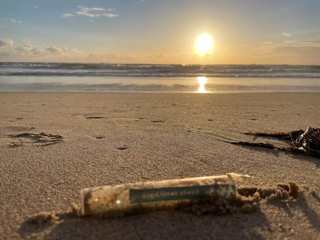 Ein kleines Röhrchen mit der Aufschrift "Gute Zeiten voraus" liegt am Sandstrand bei Sonnenuntergang, wo die Wellen schimmern und das schwache Licht der Sonne zum Nachdenken über die Bedeutung und den intuitiven Einfluss einfacher Momente einlädt.