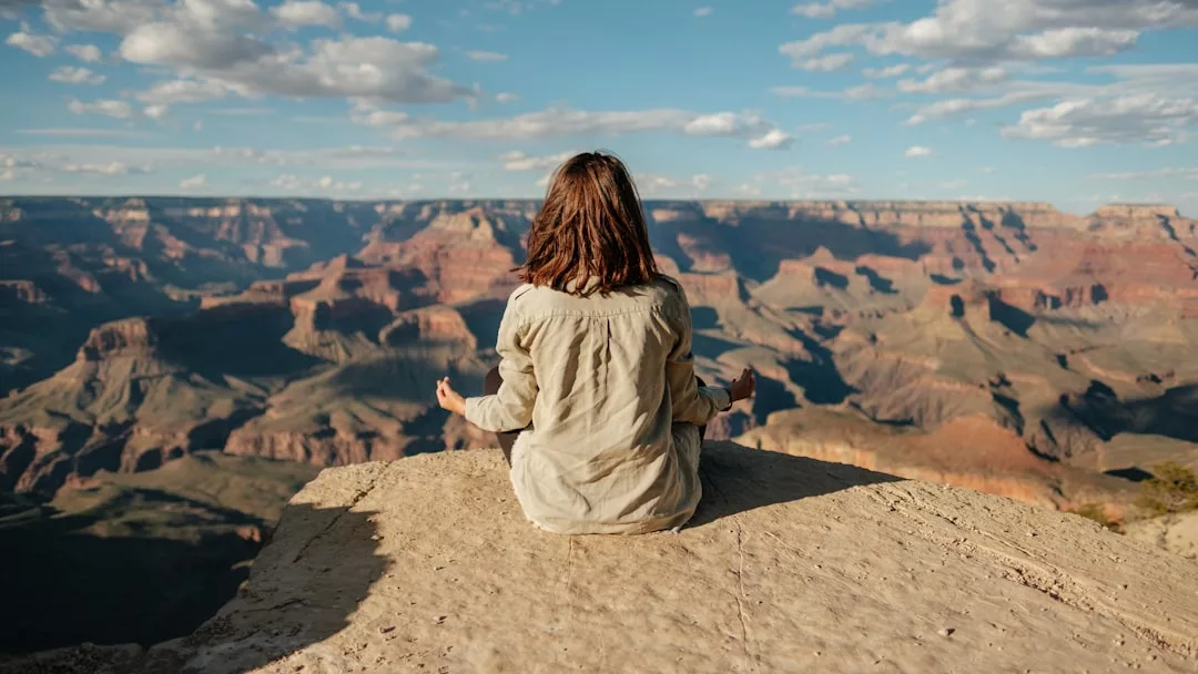 Eine Person übt sich in der Meditation, indem sie im Schneidersitz am Rande einer felsigen Klippe mit Blick auf den Grand Canyon unter einem teilweise bewölkten Himmel sitzt und inmitten der Erhabenheit der Natur innere Ruhe findet.