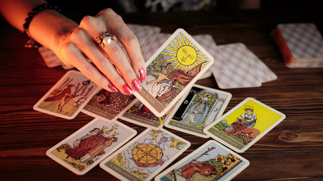 Eine Person mit lackierten Nägeln legt Tarotkarten auf einem Holztisch aus, hält die Sonnenkarte unter mehreren anderen hoch und sucht Selbsterkenntnis durch jedes Ziehen.