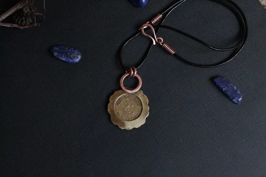 Ein runder Amulett-Anhänger aus Messing mit gezackten Rändern an einer schwarzen Kordel, mit eingraviertem Text, der auf einer dunklen Oberfläche mit blauen Steinakzenten dargestellt ist.