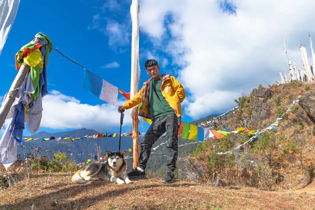 Eine Person, die auf einem Hügel steht und einen Hund an der Leine hält, bunte Gebetsfahnen flattern in der Nähe, mit Bergen und blauem Himmel im Hintergrund - in diesem ruhigen Moment denkt sie über Karma und den Einfluss des Lebens nach.