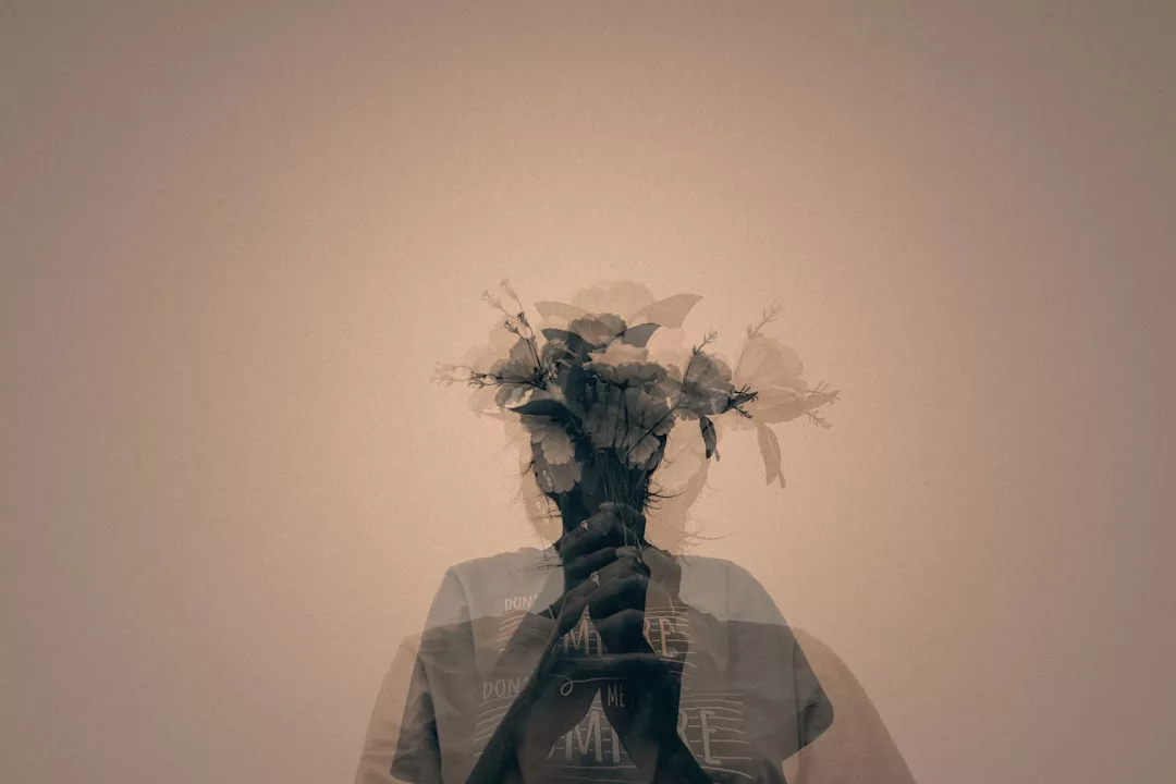 Eine Doppelbelichtung zeigt eine Person, die einen Blumenstrauß in der Hand hält, wobei die Blumen mit dem Kopf und dem Oberkörper der Person vor einem schlichten Hintergrund verschmelzen und ein Gefühl von Mystik und Verbindung zu einer spirituellen Dimension hervorrufen.