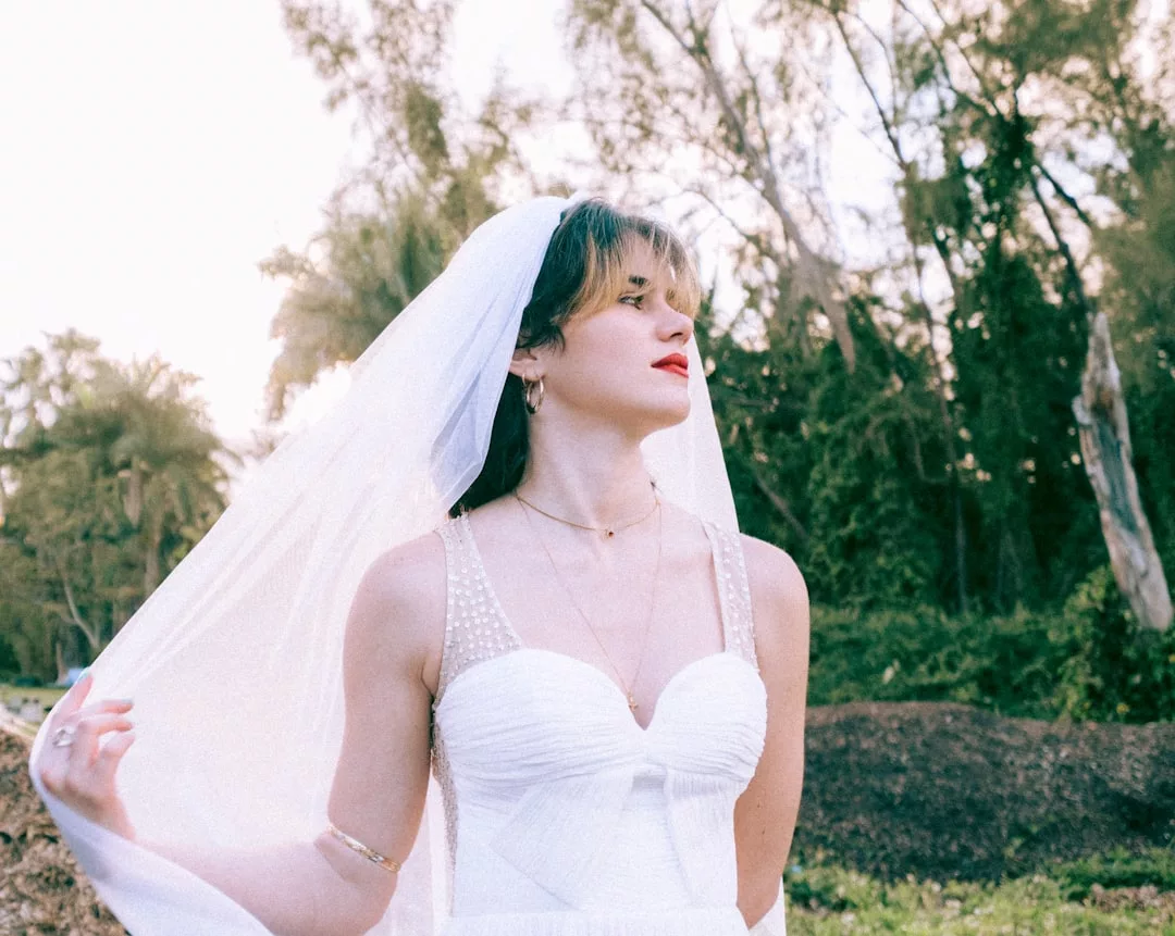 Eine Frau in weißem Hochzeitskleid und Schleier steht im Freien und umarmt den Geist des New Age, während sie mit einer Hand ihren Schleier hält, umgeben von Bäumen und üppigem Grün im Hintergrund.